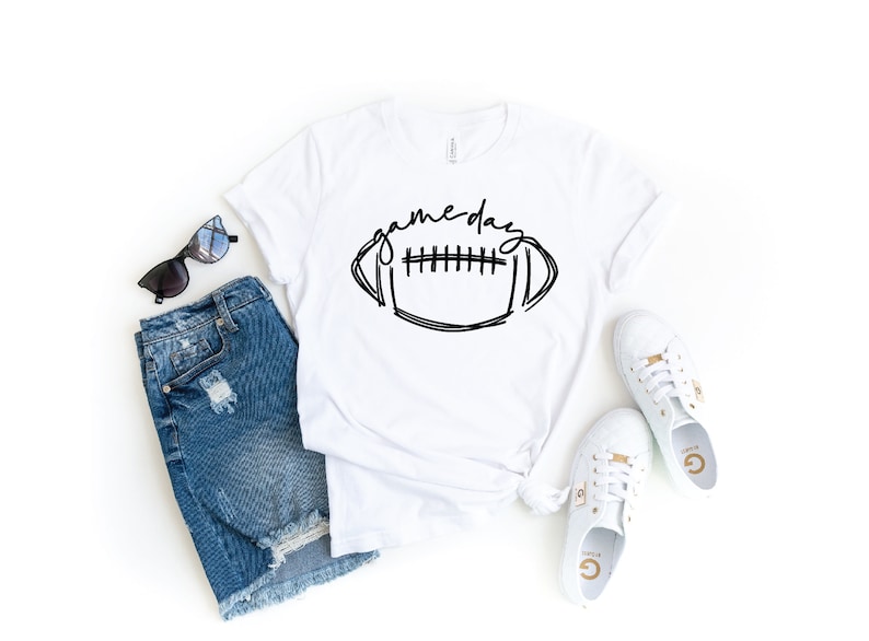 May include: White t-shirt featuring a black and white illustration of a football and the text "game day". The shirt is styled with denim shorts, sunglasses, and white trainers.