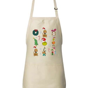 Op de afbeelding: Een beige schort met kerst-thema cartoonfiguren en elementen. Het ontwerp omvat een groene krans, een kerstboom, honden met kerstmutsen en de Grinch. De schort heeft een zak en een verstelbare nekband.