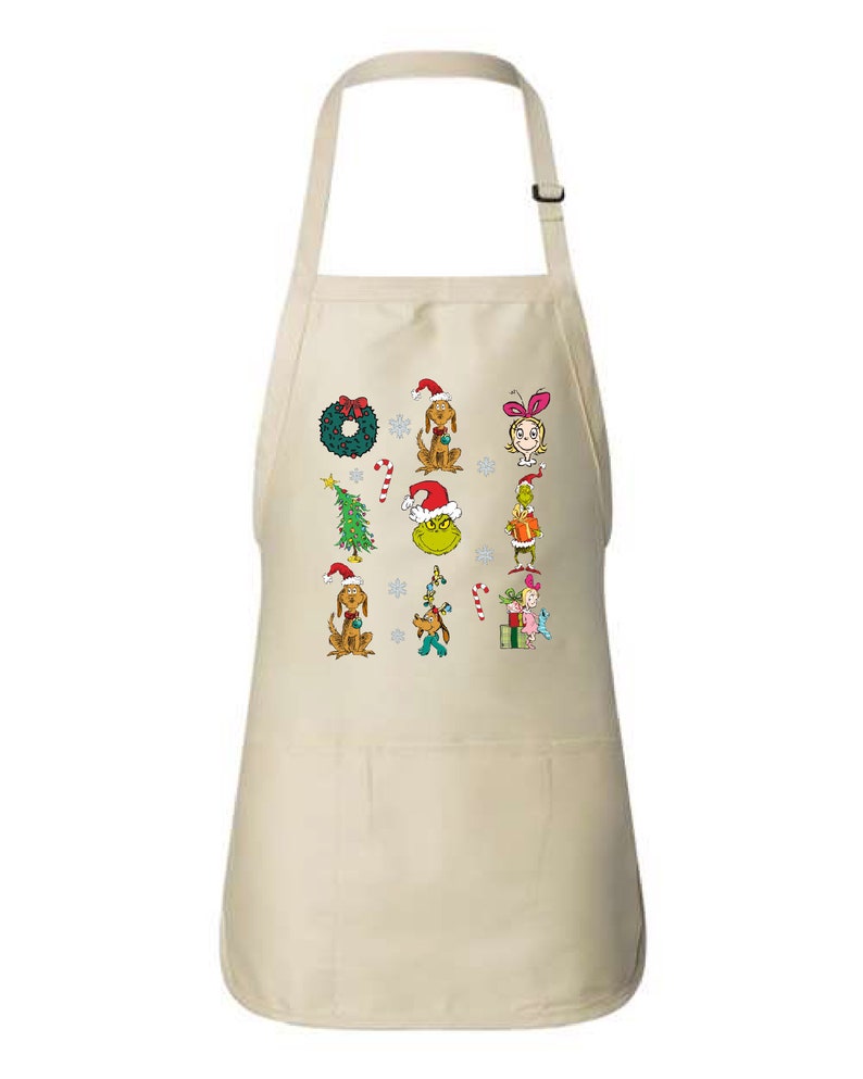 Op de afbeelding: Een beige schort met kerst-thema cartoonfiguren en elementen. Het ontwerp omvat een groene krans, een kerstboom, honden met kerstmutsen en de Grinch. De schort heeft een zak en een verstelbare nekband.