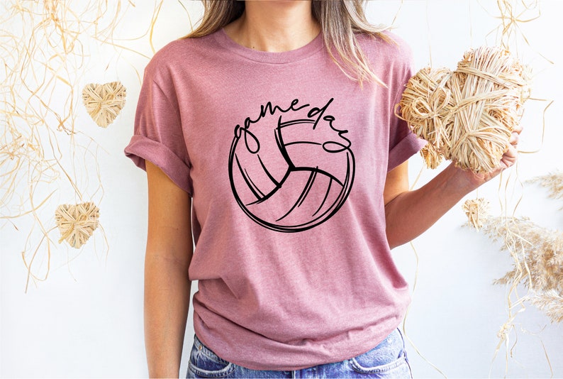 K&ouml;nnte beinhalten: Ein pinkfarbenes T-Shirt mit einer schwarzen Volleyballgrafik und dem Text "game day" um den Ball herum.