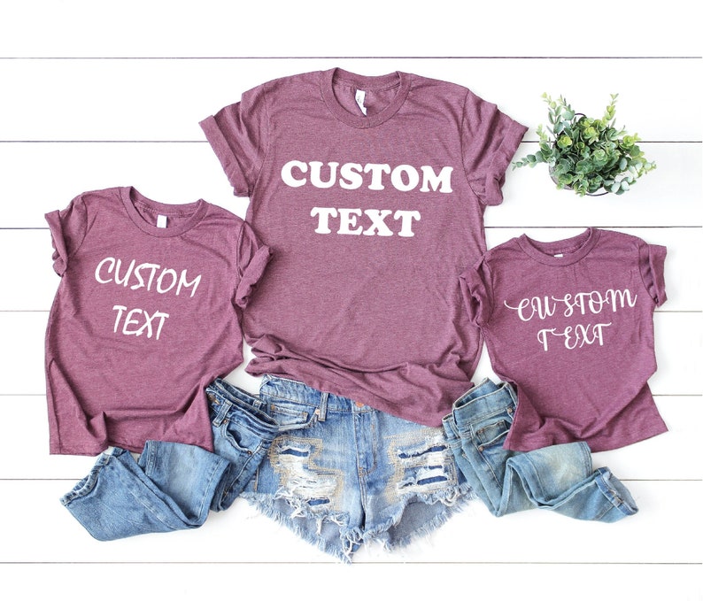 Pode incluir: Tr&ecirc;s camisetas marrons com o texto "CUSTOM TEXT" em branco. As camisetas est&atilde;o dispostas em fila, com a camiseta maior no meio e a menor &agrave; direita. As camisetas est&atilde;o sobre um fundo de madeira branco.