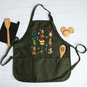 Op de afbeelding: Donkergroene schort met geborduurde kerstfiguren en symbolen. Voorzien van twee voorzakken, een houten lepel en een zwarte pannenlap. Er zijn ook chocoladekoekjes zichtbaar.