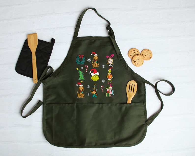Op de afbeelding: Donkergroene schort met geborduurde kerstfiguren en symbolen. Voorzien van twee voorzakken, een houten lepel en een zwarte pannenlap. Er zijn ook chocoladekoekjes zichtbaar.