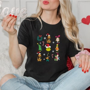 Może przedstawiać: Czarny T-shirt z grafiką świąteczną przedstawiającą postacie z Grincha i innych postaci o tematyce świątecznej. Grafika zawiera wieniec, choinkę, laski cukrowe i płatki śniegu.