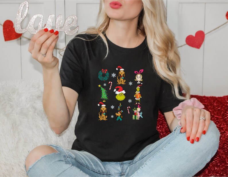 Może przedstawiać: Czarny T-shirt z grafiką świąteczną przedstawiającą postacie z Grincha i innych postaci o tematyce świątecznej. Grafika zawiera wieniec, choinkę, laski cukrowe i płatki śniegu.