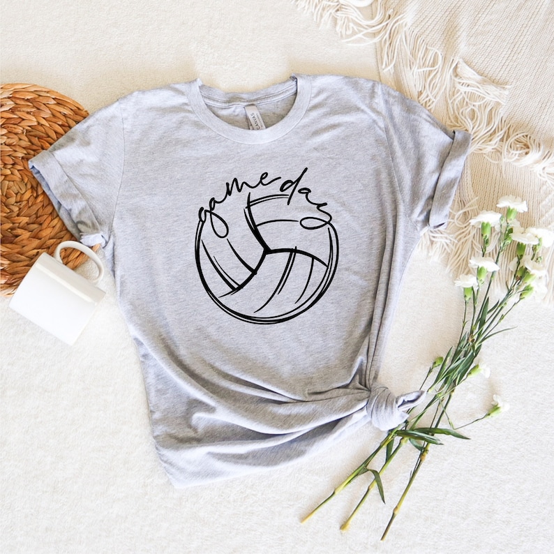 K&ouml;nnte beinhalten: Ein graues T-Shirt mit einer schwarzen Volleyballgrafik und dem Text "game day" in einer Schriftart.