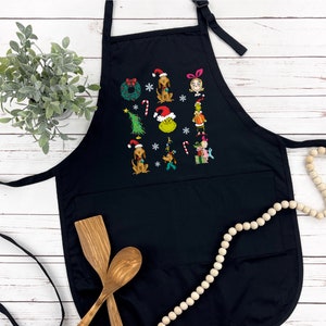 Op de afbeelding: Zwarte schort met geborduurde kerstfiguren, waaronder een hond met een kerstmuts en een groene Grinch. De schort heeft een grote voorzak en een verstelbare nekband. Houten lepels en een decoratieve slinger op de achtergrond.
