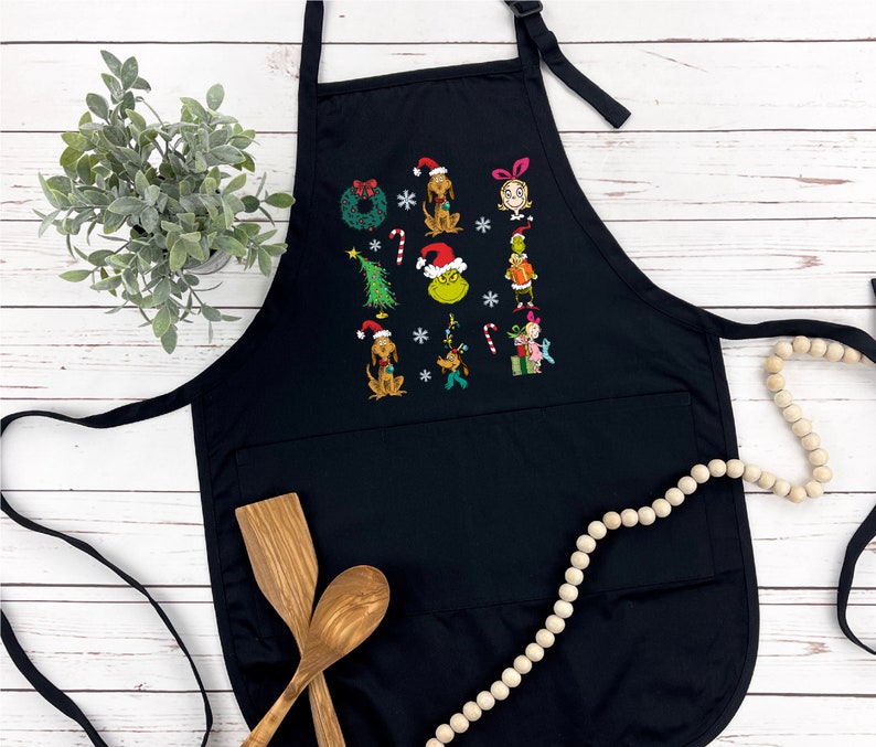 Op de afbeelding: Zwarte schort met geborduurde kerstfiguren, waaronder een hond met een kerstmuts en een groene Grinch. De schort heeft een grote voorzak en een verstelbare nekband. Houten lepels en een decoratieve slinger op de achtergrond.
