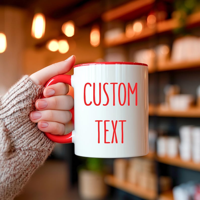 May include: White ceramic mug with a red interior and handle. The mug features the words "CUSTOM TEXT" in red lettering. The mug is held by a person wearing a beige sweater.