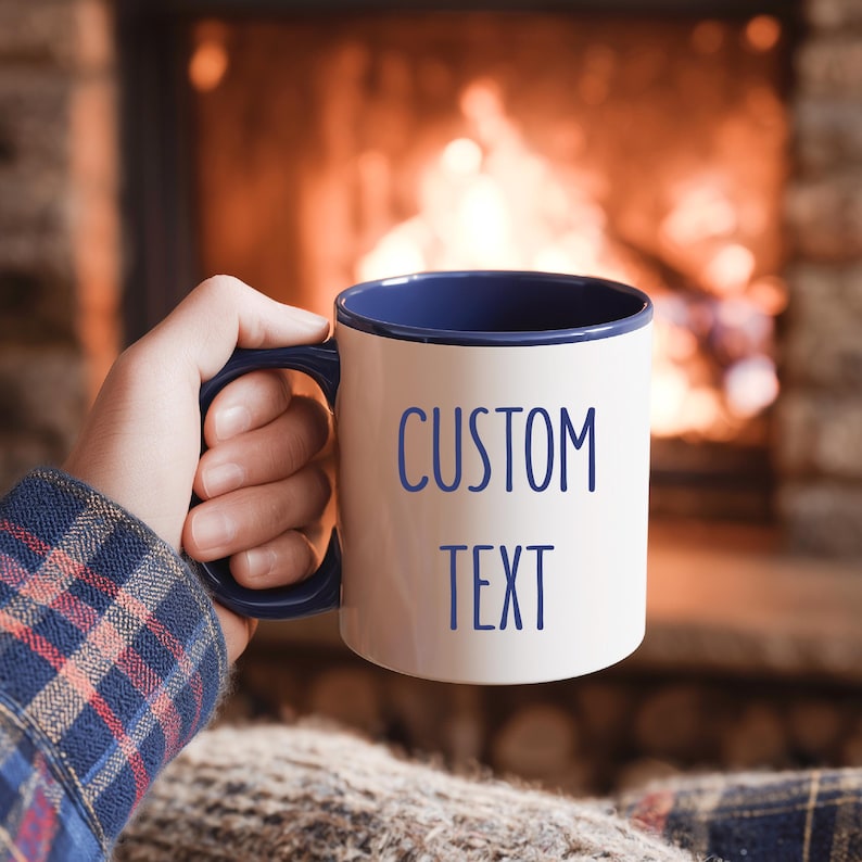 May include: A white ceramic mug with a blue interior and handle, featuring the words "CUSTOM TEXT" in blue. The mug is held by a person, with a cozy fireplace in the background.