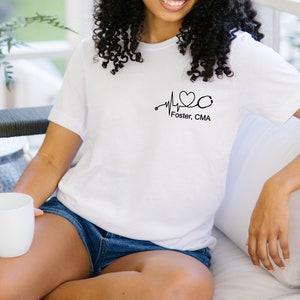 K&ouml;nnte beinhalten: Wei&szlig;es T-Shirt mit einer schwarzen Herzschlaggrafik, einem Stethoskop und dem Text "Foster, CMA".