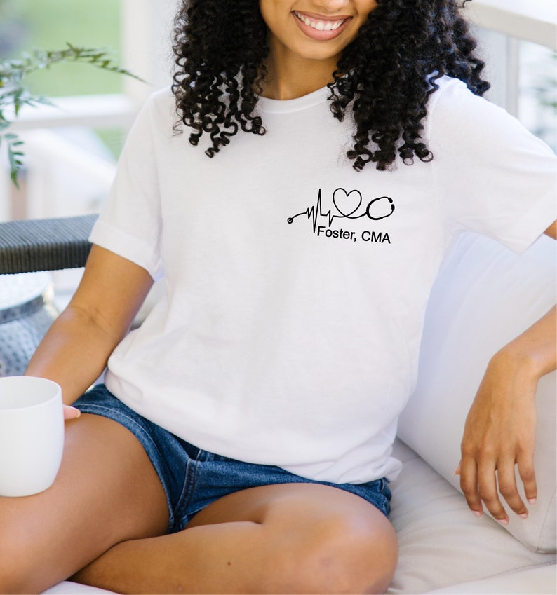 K&ouml;nnte beinhalten: Wei&szlig;es T-Shirt mit einer schwarzen Herzschlaggrafik, einem Stethoskop und dem Text "Foster, CMA".
