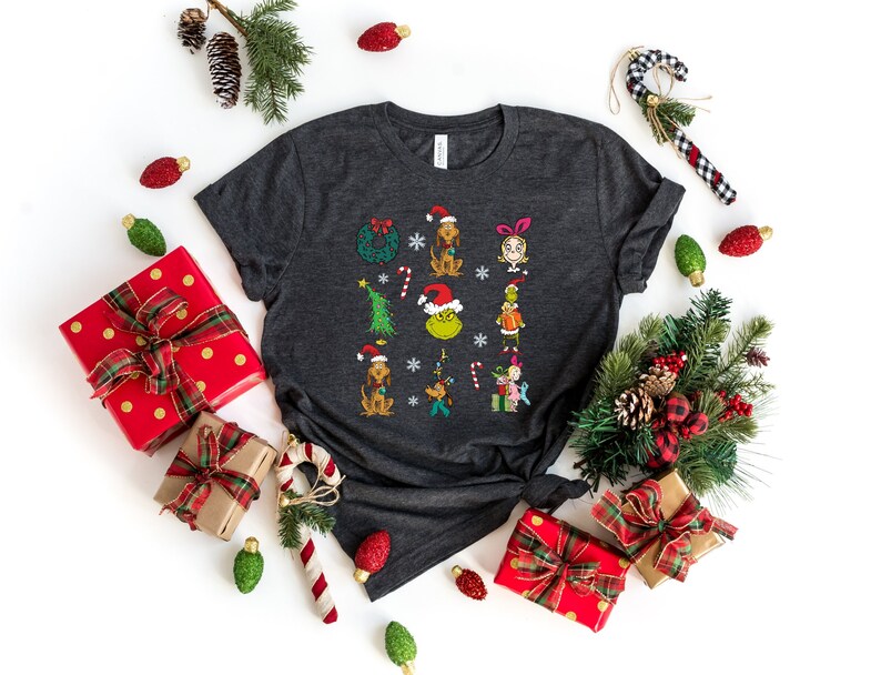 Może przedstawiać: Szary t-shirt z nadrukiem świątecznym przedstawiającym postacie z Grincha i inne symbole bożonarodzeniowe. Koszulka jest otoczona świątecznymi ozdobami, w tym ozdobami choinkowymi, laskami cukrowymi i pudełkami na prezenty.