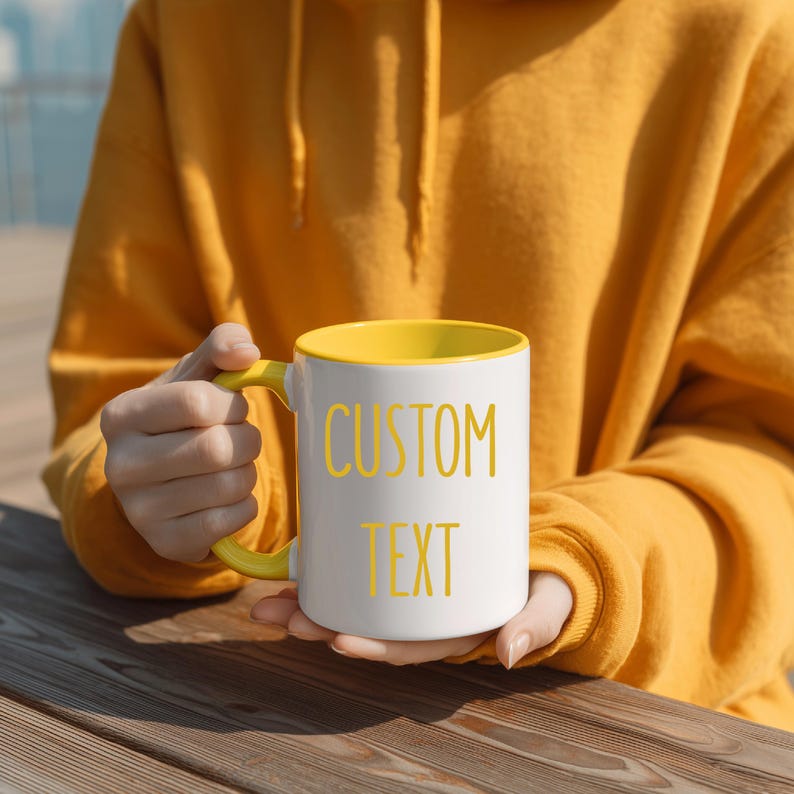 May include: White ceramic mug with a yellow interior, handle, and the words "CUSTOM TEXT" in yellow. The mug is held by a person wearing a yellow hoodie. The mug is on a wooden surface.