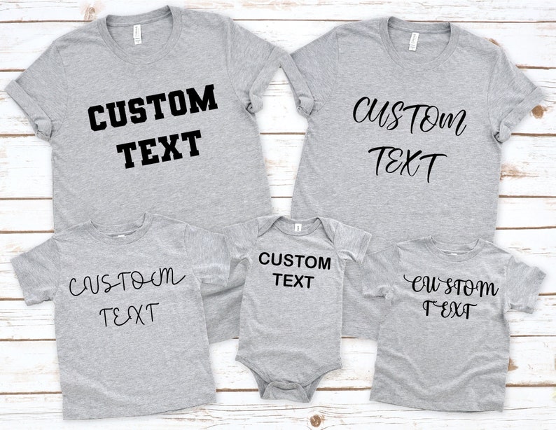 Pode incluir: Quatro t-shirts cinzas com o texto "CUSTOM TEXT" em preto. As t-shirts s&atilde;o de tamanhos e estilos diferentes, uma delas &eacute; um macac&atilde;o para beb&ecirc;.