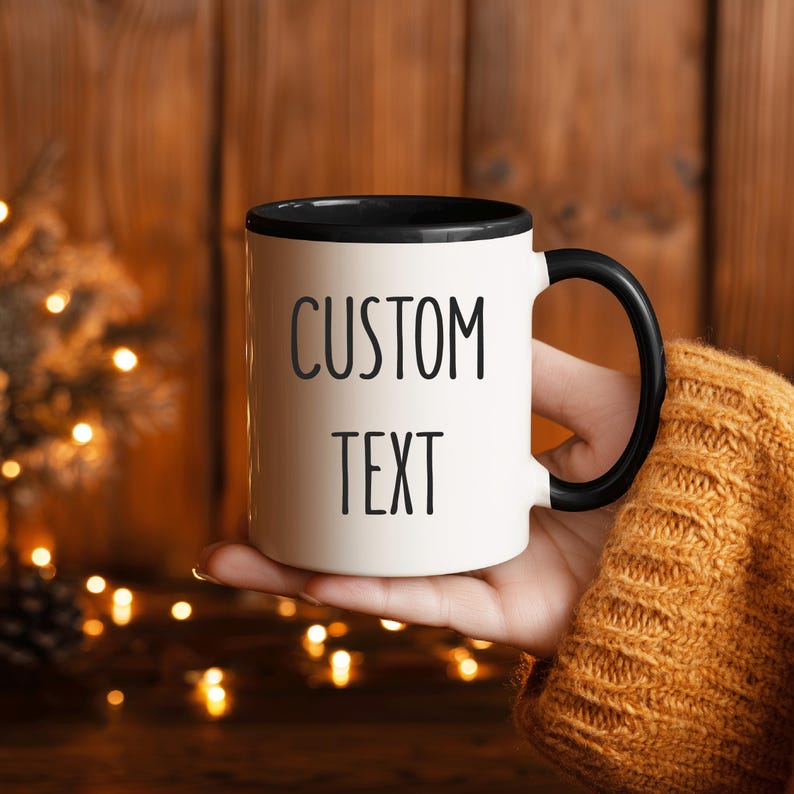 May include: White ceramic mug with a black rim and handle, featuring the words "CUSTOM TEXT" in black capital letters. The mug is held against a blurred background of warm-toned wood and twinkling lights.