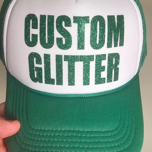May include: A green and white trucker hat with the words "CUSTOM GLITTER" in large, glittery green letters across the front. The hat has a green brim and mesh backing. The text "GREEN GLITTER" is at the bottom.