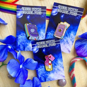 Peut inclure: Trois épingles en émail sur des cartes avec un fond violet et un design de ciel étoilé. Les épingles sont un livre violet avec le texte "Gay Tarot" sur la couverture, un livre violet avec le texte "Trans Tarot" sur la couverture, et une bouteille violette avec une étiquette qui dit "Spell Bottle". Les cartes ont le texte "Queer Witch Enamel Pins" sur elles.