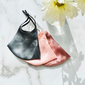 May include: Three silk face masks. One is dark grey, one is light pink, and one is a darker shade of pink. The masks are folded and stacked on top of each other.