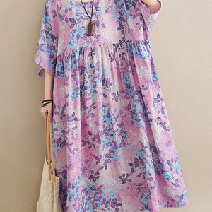 May include: A loose-fitting midi dress featuring a vibrant floral pattern in shades of purple, lavender, and bright blue. The garment has a round neckline, short sleeves, and a gathered waistline that creates a relaxed, flowing silhouette.