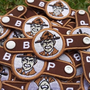 May include: A collection of brown and white embroidered patches. Each patch features a circular design with a portrait of a man in a hat and a brown tab with the letter "B" or "P" and a white button.