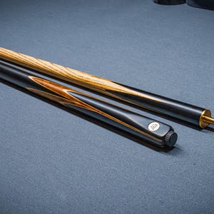 Op de afbeelding: Een tweedelige snookerkeu met een lichtbruine essenhouten schacht met een opvallende houtnerf en een zwart achterstuk met decoratieve houten inlegstukken. De keu is voorzien van een messing verbinding, een zwarte rubberen dop en een wit logo.