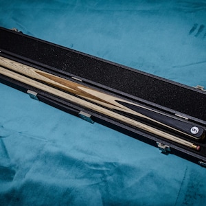 May include: A two-piece wooden snooker cue with a light tan wood grain shaft and a dark brown butt section featuring a black splice design. The cue is housed in a rectangular black hard case with a black foam interior and metal latches.