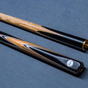 May include: A two-piece snooker cue featuring a light brown wood grain shaft with black spliced accents. The butt section displays a decorative pattern of black, blue, and light wood veneers, finished with a brass joint and a circular white logo inlay.