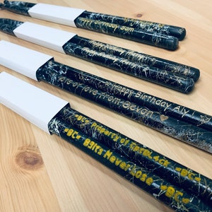 May include: A set of black and gold drumsticks with personalized messages. The drumsticks are decorated with a white marble pattern and have gold lettering. The messages include "Happy Birthday Aly", "Lots of love from Devon", and "BC - Beats Never Lose - BC".