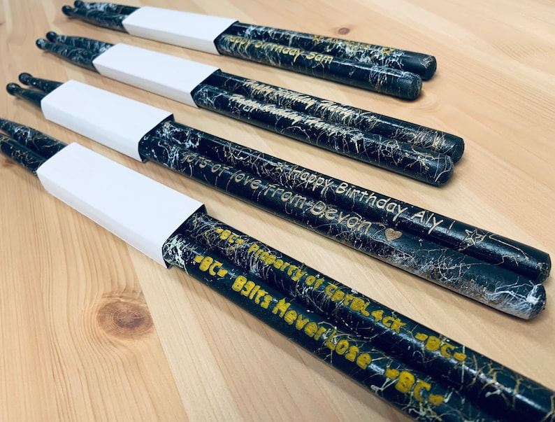 May include: A set of black and gold drumsticks with personalized messages. The drumsticks are decorated with a white marble pattern and have gold lettering. The messages include "Happy Birthday Aly", "Lots of love from Devon", and "BC - Beats Never Lose - BC".