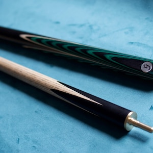 May include: A two-piece pool cue featuring a light wood shaft with black points and a dark butt section with green and black wood grain patterns. The butt end displays a circular silver logo with a stylized letter, and the joint has a brass pin.