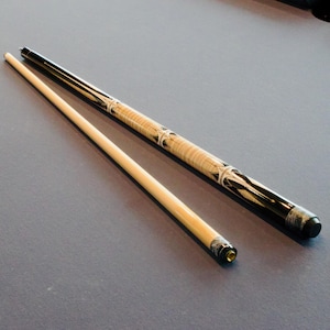May include: A two-piece pool cue with a light wood shaft and a black butt. The butt features star-shaped inlays and a light wood section. The cue is lying on a grey surface.