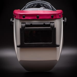 May include: A welding helmet with a white body, a black face shield, and a red top. The helmet has a dark gray head support and black adjustment knobs. The face shield is rectangular and dark.