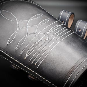 May include: Black leather arm guard with white stitching in a geometric pattern. The guard has multiple straps with black stitching.