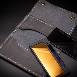 May include: A gray leather wallet with white stitching and two black welding lens filters. One filter has a gold reflective surface, and the other is black with the text "LINCOLN SUPERVISIBILITY-NO.10-H".