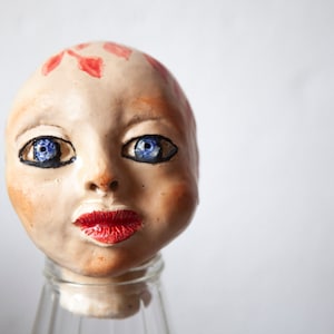 May include: A ceramic doll head with painted red details on the forehead, blue eyes, and red lips. The head is sitting on a clear glass jar.