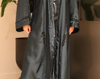 Op de afbeelding: Een lange, donkerblauwe abaya met zwarte kralenversieringen. De abaya heeft lange mouwen en is aan de voorkant open, waardoor een zwarte top en jeans zichtbaar zijn. Een zwarte handtas met een gouden ketting is zichtbaar.