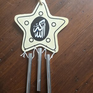 Peut inclure: Une décoration suspendue en bois en forme d'étoile avec une surface crème, des contours noirs et quatre petites découpes circulaires. Un cercle noir central présente une calligraphie arabe blanche. Trois tubes métalliques argentés pendent.