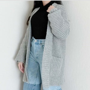 Pode incluir: Um casaco de malha cinzento com textura canelada e dois bolsos. O casaco está aberto e usado sobre uma camisa preta de gola alta e calças de ganga azuis.