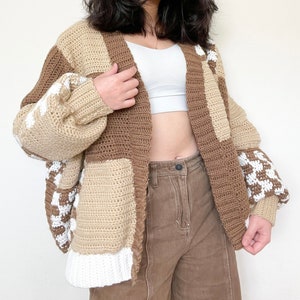 May include: A brown and white crocheted cardigan with a patchwork design. The cardigan has a loose fit and long sleeves.