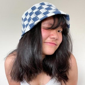 Op de afbeelding: Een persoon draagt ​​een blauwe en witte geruite bucket hat. De hoed is gemaakt van zacht, gecrocheteerd materiaal.