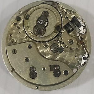 Puede incluir: Primer plano de un movimiento de reloj antiguo, que muestra intrincados componentes mec&aacute;nicos. Se ven los engranajes, muelles y volante plateados. El mecanismo del reloj tiene forma circular con peque&ntilde;os tornillos y marcas.