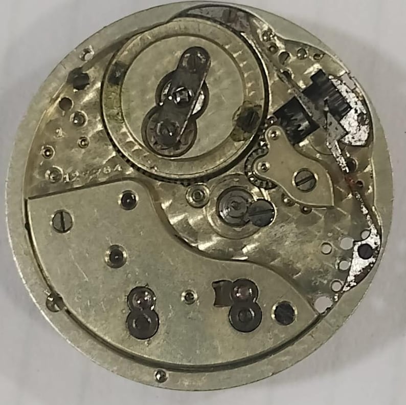 Puede incluir: Primer plano de un movimiento de reloj antiguo, que muestra intrincados componentes mec&aacute;nicos. Se ven los engranajes, muelles y volante plateados. El mecanismo del reloj tiene forma circular con peque&ntilde;os tornillos y marcas.