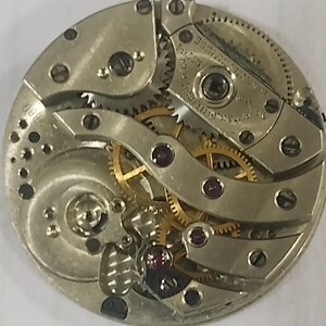 Puede incluir: Primer plano de un movimiento de reloj antiguo. El intrincado mecanismo presenta numerosos engranajes, joyas y componentes met&aacute;licos. El movimiento del reloj es circular, con un acabado plateado y engranajes dorados. La imagen destaca la precisi&oacute;n y la artesan&iacute;a del reloj.