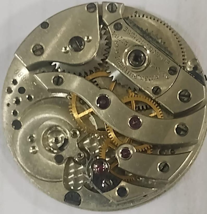 Puede incluir: Primer plano de un movimiento de reloj antiguo. El intrincado mecanismo presenta numerosos engranajes, joyas y componentes met&aacute;licos. El movimiento del reloj es circular, con un acabado plateado y engranajes dorados. La imagen destaca la precisi&oacute;n y la artesan&iacute;a del reloj.