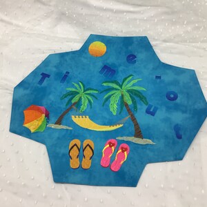 Könnte beinhalten: Ein blauer, sechseckiger Wandbehang aus Stoff mit dem Wort "Time Out", das in bunten Buchstaben gestickt ist. Das Design beinhaltet Palmen, eine Hängematte, einen Sonnenschirm, Flip-Flops und eine Sonne.