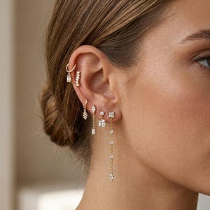 May include: A collection of gold-toned earrings worn in a single ear, featuring a small hoop with a rectangular stone, a bar stud, a hoop with a dangling marquise stone, a stud with a square stone, and a long chain earring with multiple stones.
