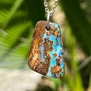 Puede incluir: Collar con colgante de piedra natural y cadena de plata. El colgante presenta un patrón único de tonos marrones, azules y verdes. La piedra, de forma irregular, parece pulida. El collar se muestra sobre un fondo verde borroso.