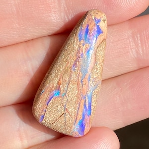 Op de afbeelding: Een bruine en beige opaal hanger met blauwe en paarse iriserende flitsen. De opaal heeft een driehoekige vorm met een ruwe, natuurlijke textuur.