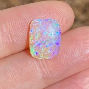 May include: A white opal cabochon with a rectangular shape and a rainbow of colours, including blue, green, and purple.